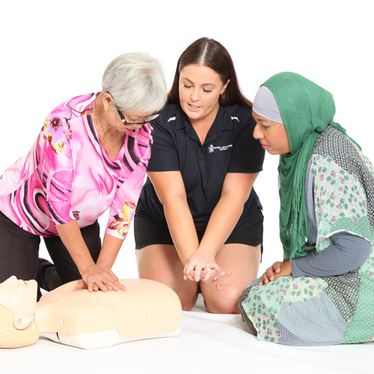 Woman wearing a royal life saving society shirt with two women on either side and a CPR dummy on the floor demonstrating CPR